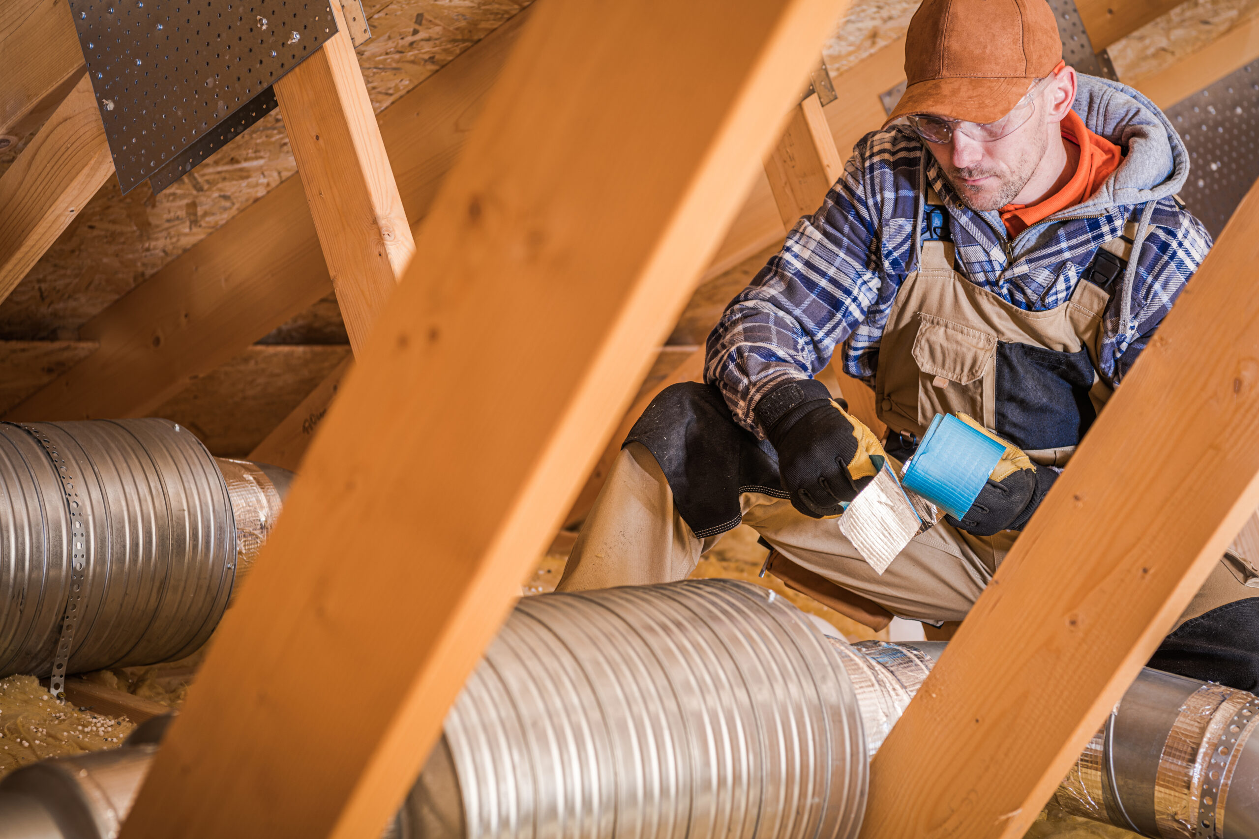 Construction Worker Wrapping HVAC Air Duct With Silver And Blue Foil Tape In Attic Of Residential Building.